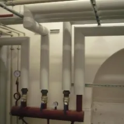 Izolowane rury pionowe z zaworami i manometrami, część instalacji w piwnicy lub kotłowni, widoczne rury pod sufitem.