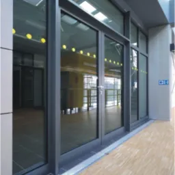 Duże, grafitowe okna aluminiowe z podwójnymi drzwiami wejściowymi, widoczne z zewnątrz budynku, z jasną podłogą imitującą drewno.