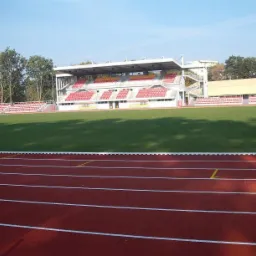 Widok og&oacute;lny stadionu z bieżnią tartanową, zieloną murawą i trybunami z czerwonymi krzesełkami w słoneczny dzień.