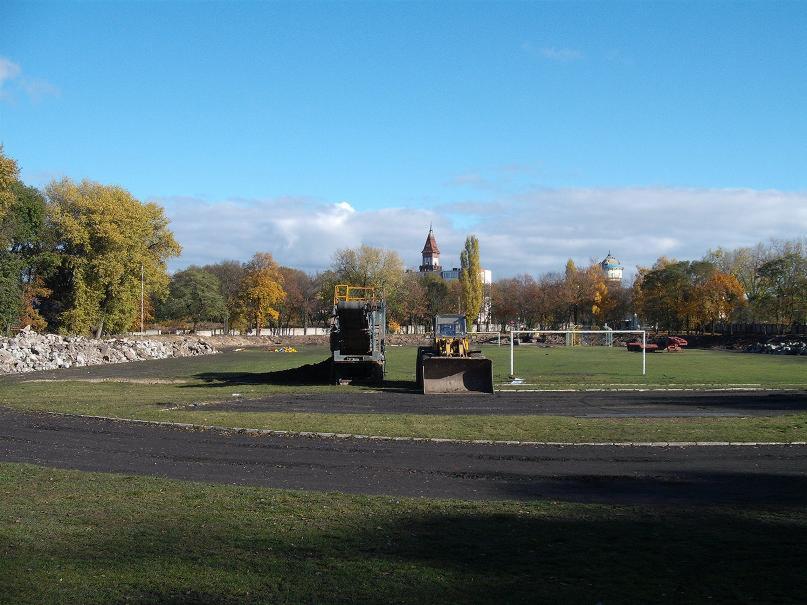 Przygotowanie terenu stadionu z użyciem ciężkiego sprzętu: spychacz i przesiewacz do ziemi, w tle budynki miejskie.