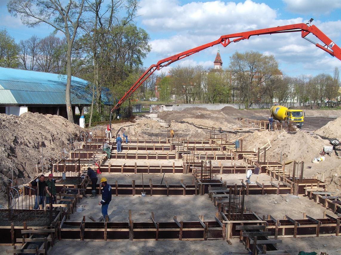 Wylewanie betonu do szalunków fundamentowych na placu budowy, widoczny dźwig z pompą do betonu i betoniarka w tle.