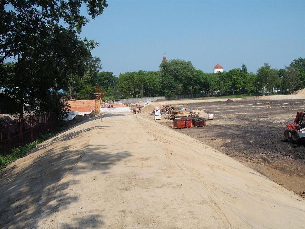 Teren budowy z widocznym fundamentem budynku w trakcie wznoszenia, ziemią wyrównaną i przygotowaną pod dalsze prace, oraz czerwonym spychaczem w prawym dolnym rogu. W tle widoczne drzewa i wieża...