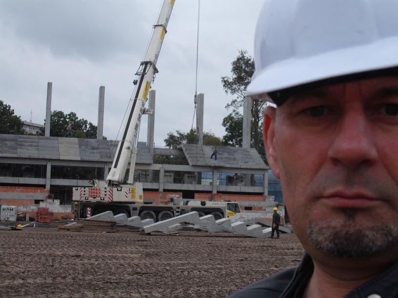 Budowa stadionu w trakcie montażu prefabrykatów, widok z perspektywy osoby w białym kasku na pierwszym planie, w tle żuraw wieżowy i pracownicy.