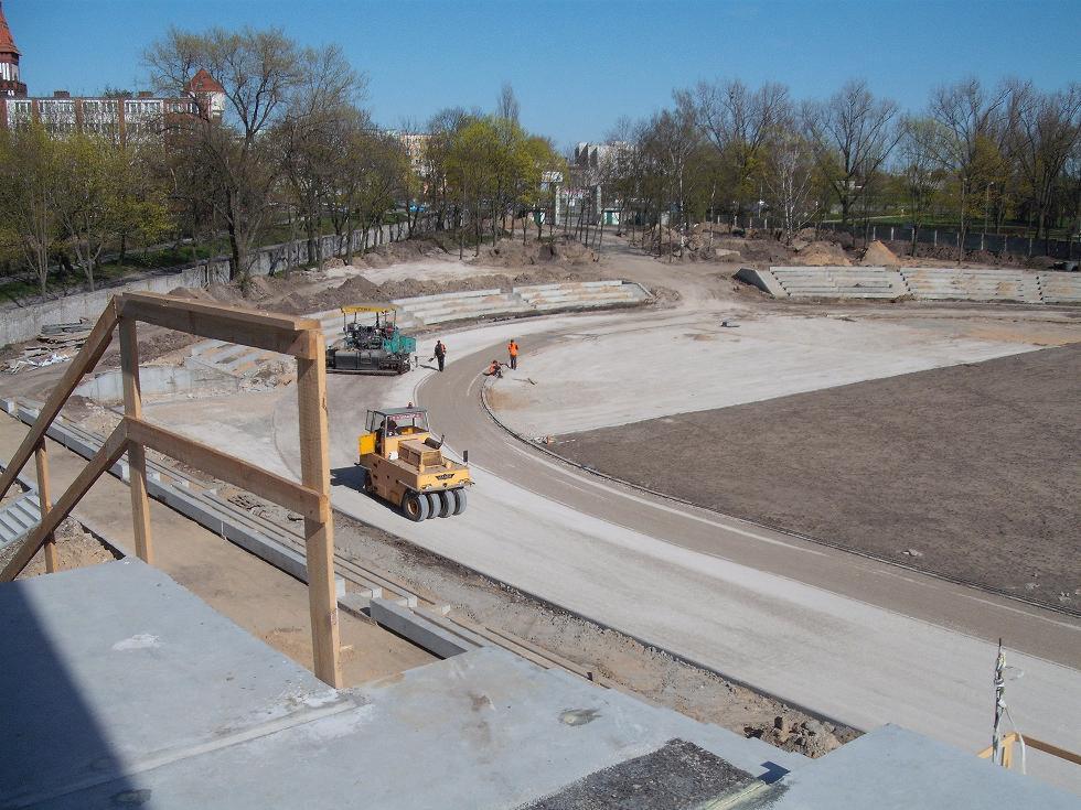 Budowa stadionu z widocznymi maszynami budowlanymi: walcem drogowym i rozściełaczem asfaltu, w tle budynki mieszkalne i drzewa.