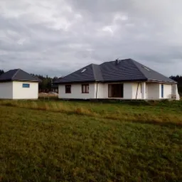 Nowo wybudowany, parterowy dom jednorodzinny z szarą dachówką i białymi ścianami, obok mały budynek gospodarczy, otoczone wysoką trawą na tle pochmurnego nieba.