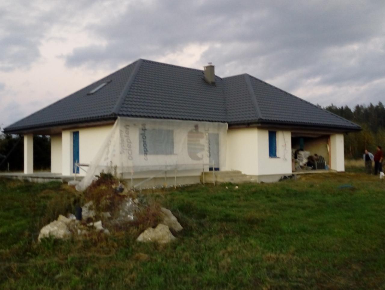 Jednopiętrowy dom z ciemnoszarą dachówką, otynkowany na biało, z niebieskimi okiennicami, częściowo osłonięty folią budowlaną, widoczny w trakcie budowy na tle pochmurnego nieba.