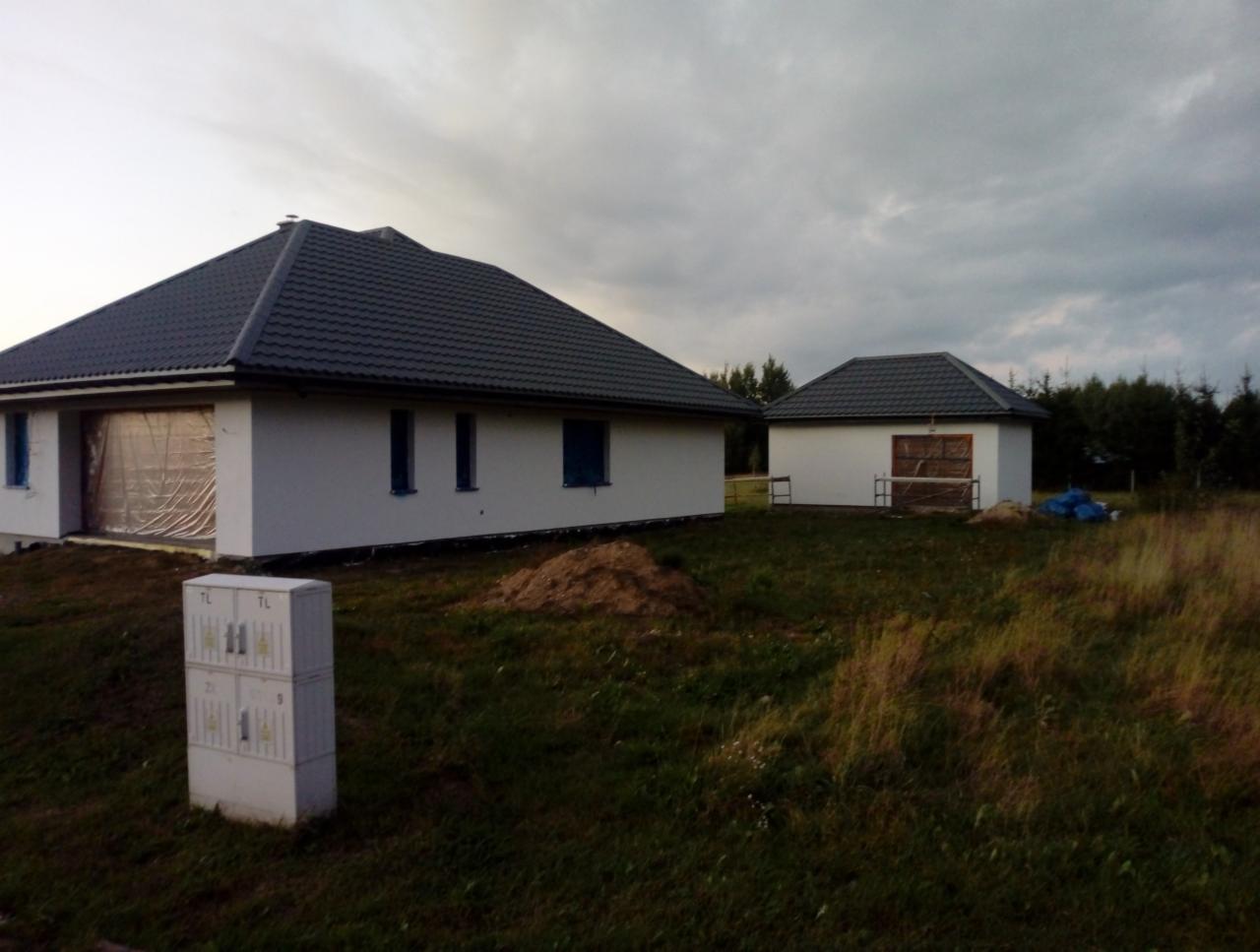 Nowo wybudowany parterowy dom z szarą dachówką i białymi ścianami, okna zabezpieczone folią, obok drugi budynek gospodarczy w podobnym stylu, widoczne trawy i pochmurne niebo.