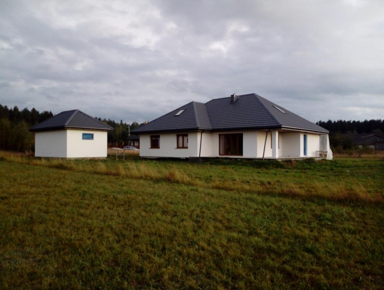 Nowo wybudowany, parterowy dom jednorodzinny z szarą dachówką i białymi ścianami, obok mały budynek gospodarczy, otoczone wysoką trawą na tle pochmurnego nieba.