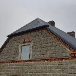 Szary dach z blachy trapezowej na murowanym domu z ceglanymi detalami, widoczne kominy i zabezpieczone okno na szczycie budynku.