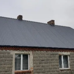 Dom z szarej cegły z nowym, ciemnoszarym dachem z blachy trapezowej, widoczne dwa kominy z czerwonej cegły i rynny w kolorze dachu.