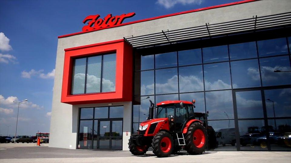 Czerwony traktor Zetor na tle nowoczesnego budynku z dużymi oknami, odbijającymi niebo i chmury.