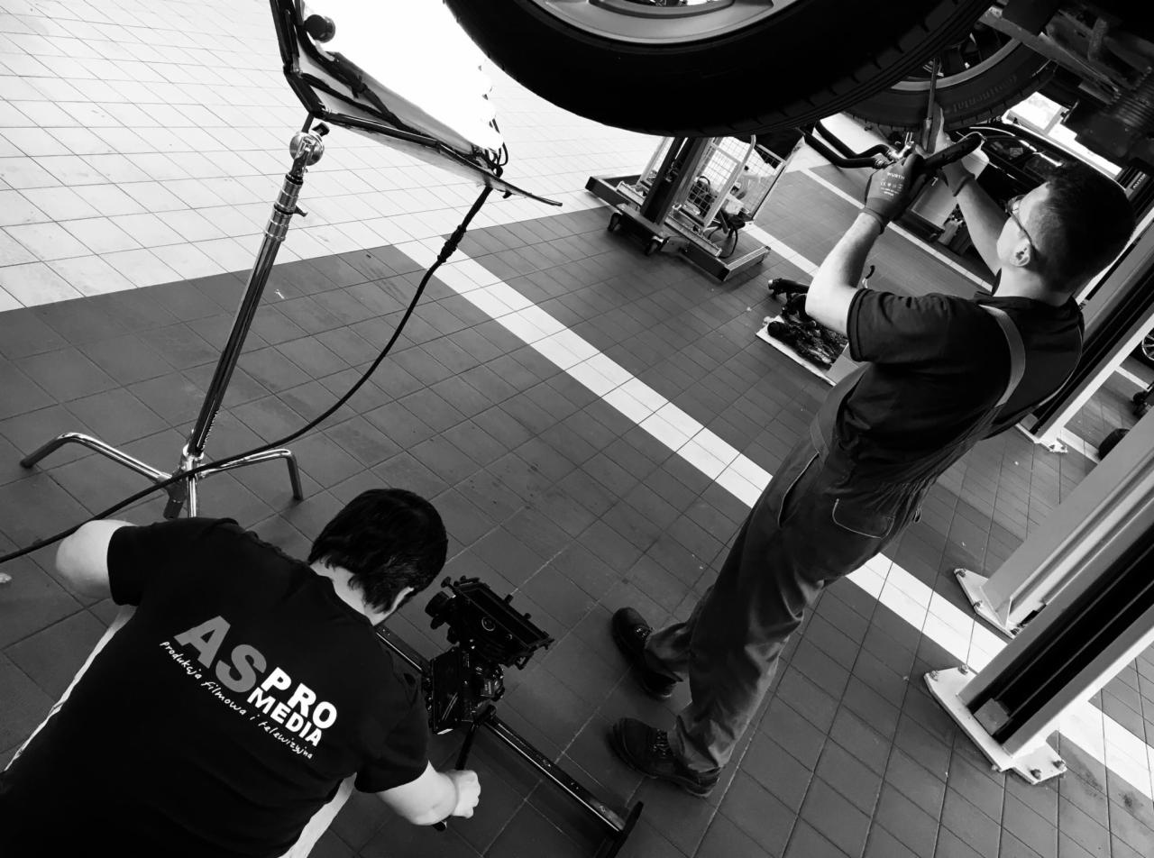 Czarno-białe zdjęcie z planu filmowego: operator kamery z logo firmy na koszulce, filmujący mechanika samochodowego pracującego pod podniesionym autem, widoczny statyw oświetleniowy.