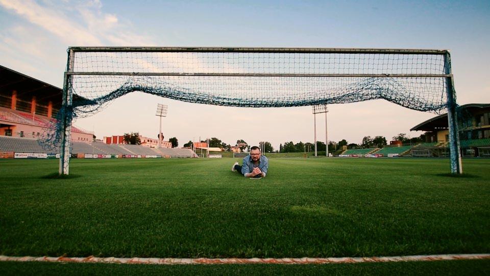 Mężczyzna w okularach leży na murawie boiska piłkarskiego, z bramką w tle, prawdopodobnie podczas kręcenia filmu.