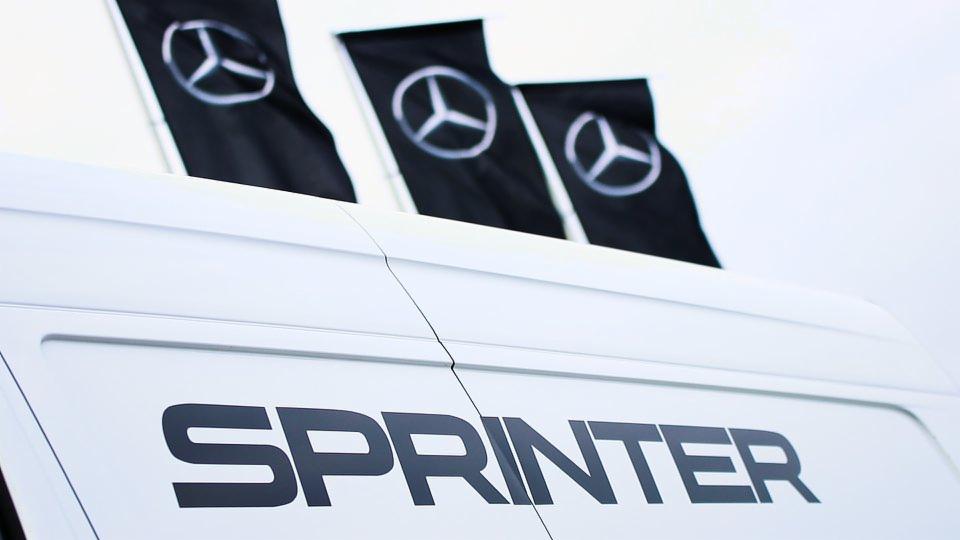 Biały samochód dostawczy marki Sprinter z trzema czarnymi flagami z logo producenta w tle.