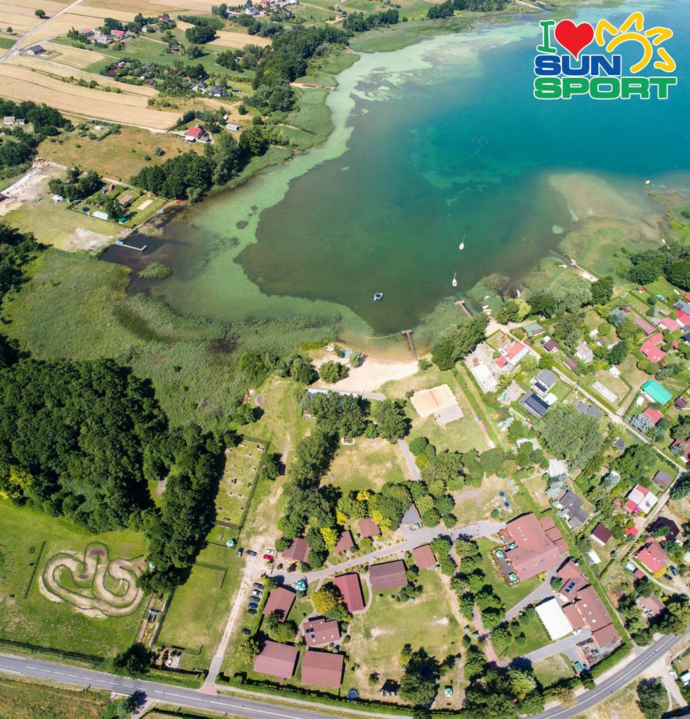 Widok z lotu ptaka na jezioro z piaszczystą plażą, otoczone zielenią i zabudowaniami. W prawym górnym rogu widoczne logo 'I love SUN Sport'.