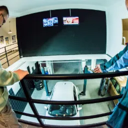 Widok z góry na warsztat samochodowy z dwoma pracownikami przy barierce, biały samochód na podnośniku oraz monitory pokazujące proces naprawy.