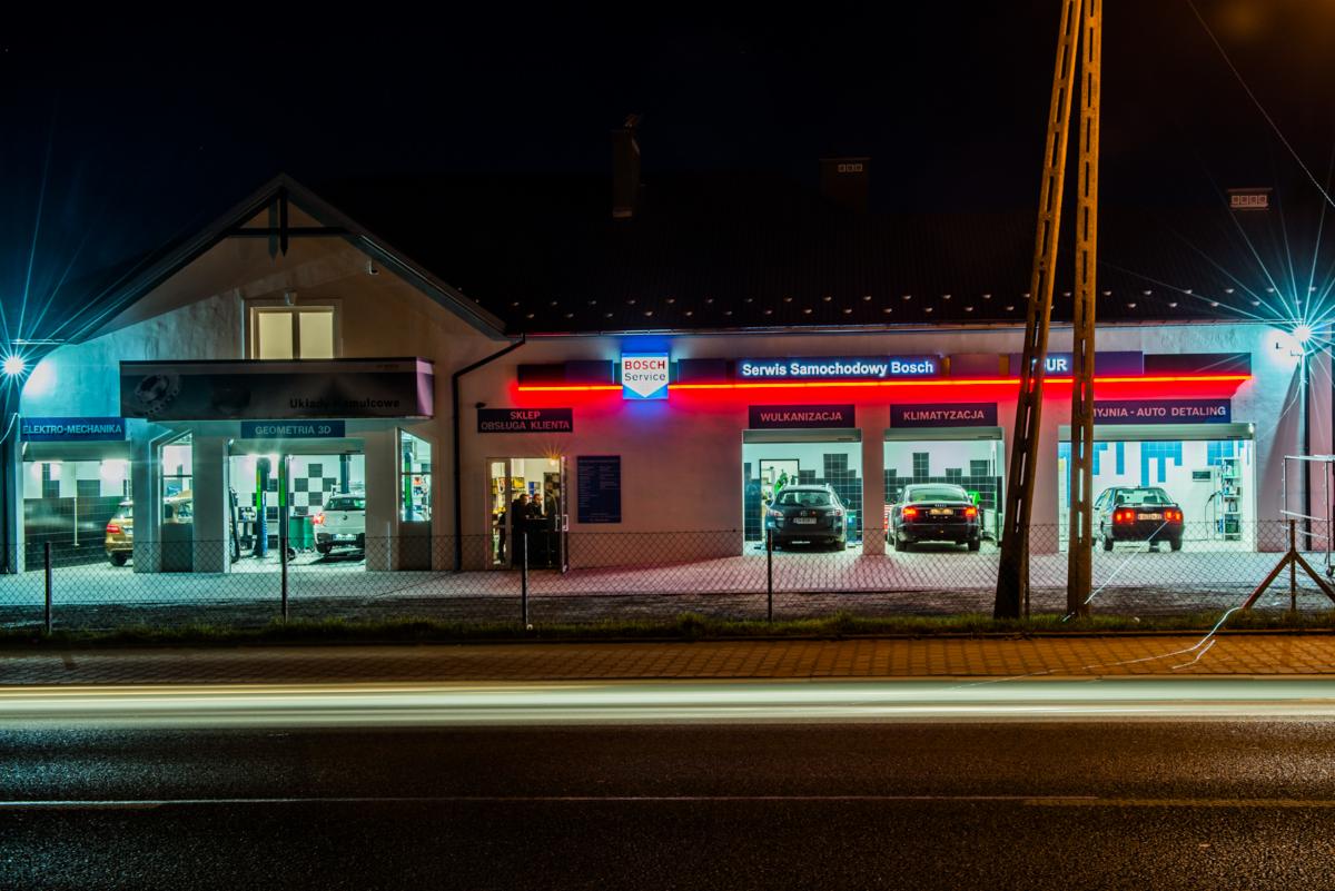 Nocny widok na fasadę serwisu samochodowego Bosch z neonowymi napisami nad stanowiskami: Elektro-Mechanika, Geometria 3D, Wulkanizacja, Klimatyzacja i Myjnia-Auto Detailing. Przed budynkiem...