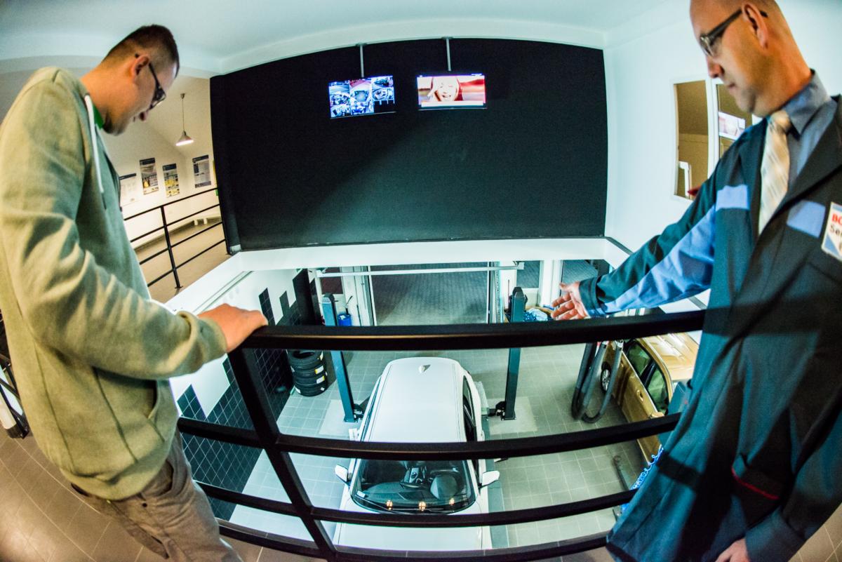 Widok z góry na warsztat samochodowy z dwoma pracownikami przy barierce, biały samochód na podnośniku oraz monitory pokazujące proces naprawy.