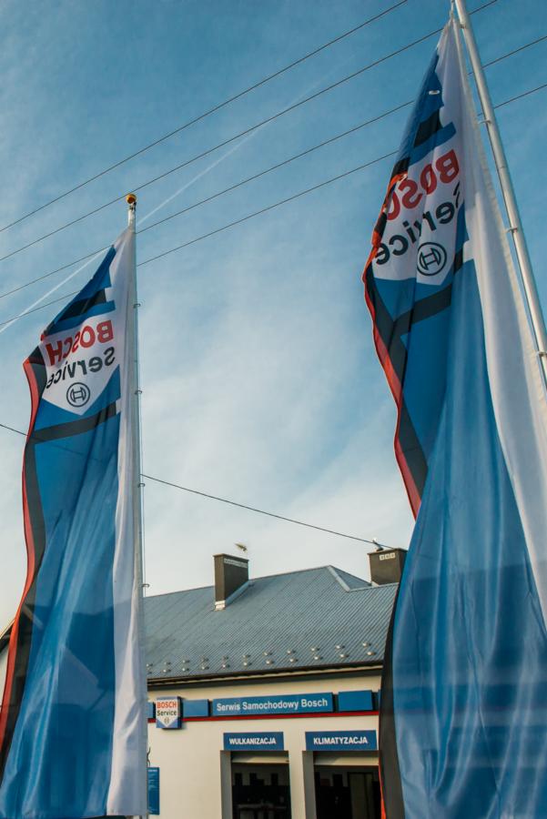 Dwa flagi z logo Bosch Service powiewające przed budynkiem serwisu samochodowego z napisem 'Serwis Samochodowy Bosch', 'Wulkanizacja', 'Klimatyzacja' na tle błękitnego nieba.
