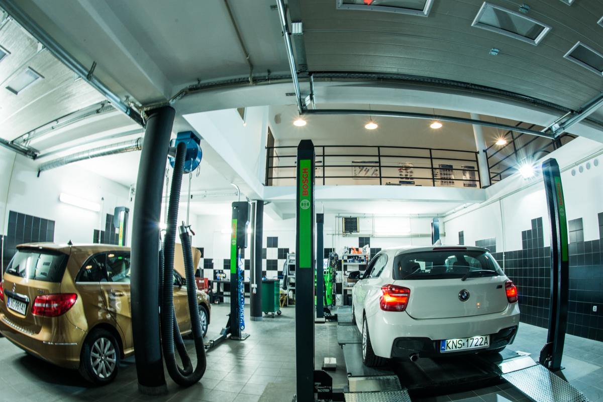 Wnętrze warsztatu samochodowego z dwoma samochodami: złoty Mercedes klasy B i białe BMW serii 1, oba na podnośnikach. W tle widoczne narzędzia i wyposażenie warsztatowe, a także antresola.