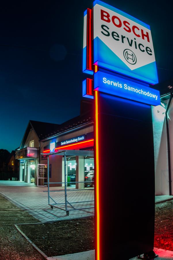 Oświetlony nocą szyld z logo Bosch Service przed budynkiem serwisu samochodowego z przeszklonymi wjazdami.