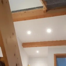 Biały sufit z drewnianymi belkami, oświetleniem punktowym i fragmentem schodów z widoczną konstrukcją nośną, ukazujący wnętrze w trakcie prac wykończeniowych.