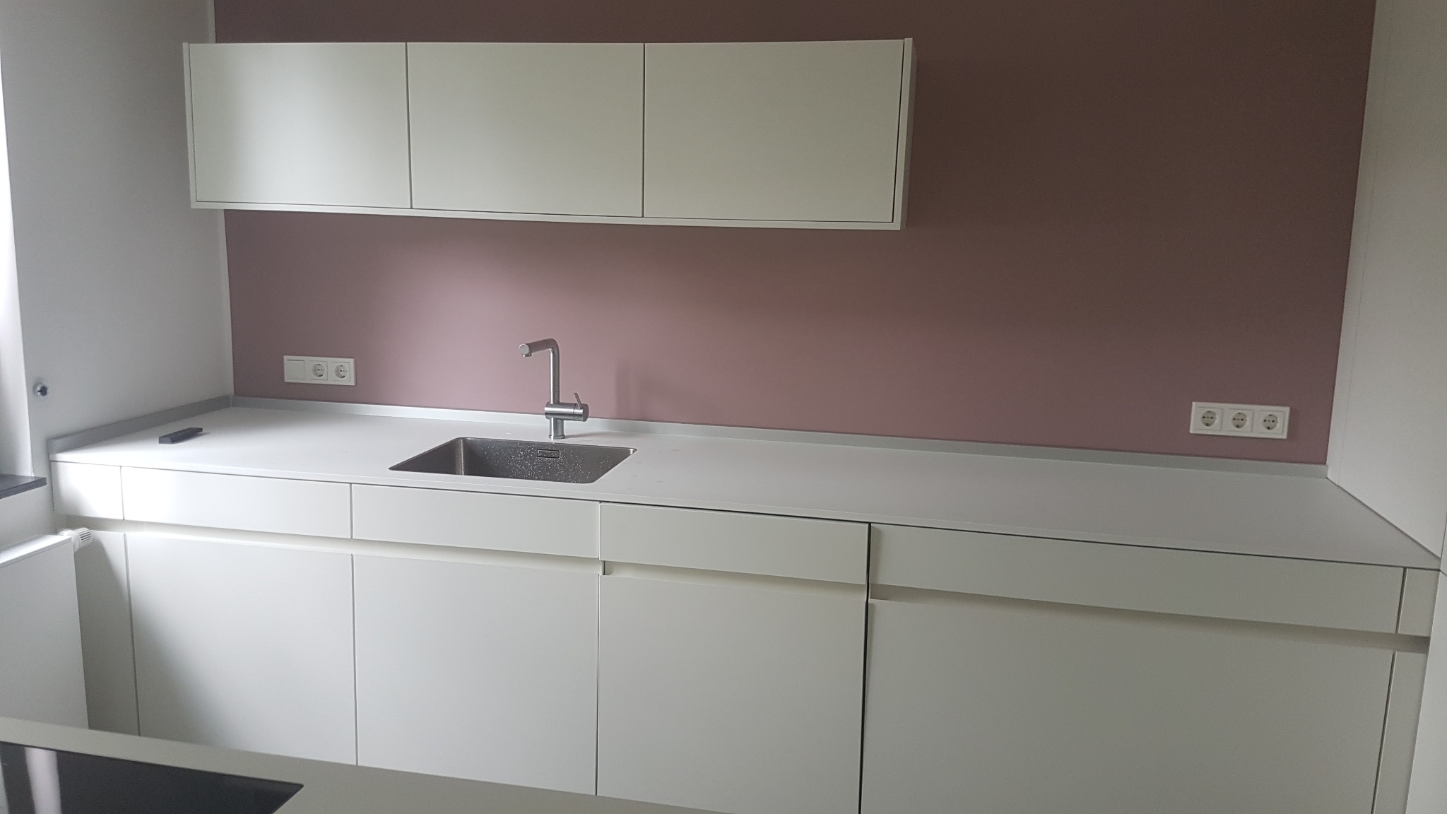 Nowoczesna kuchnia z białymi szafkami, szarym blatem i ścianą pomalowaną na kolor pudrowego różu, widoczne gniazdka elektryczne.