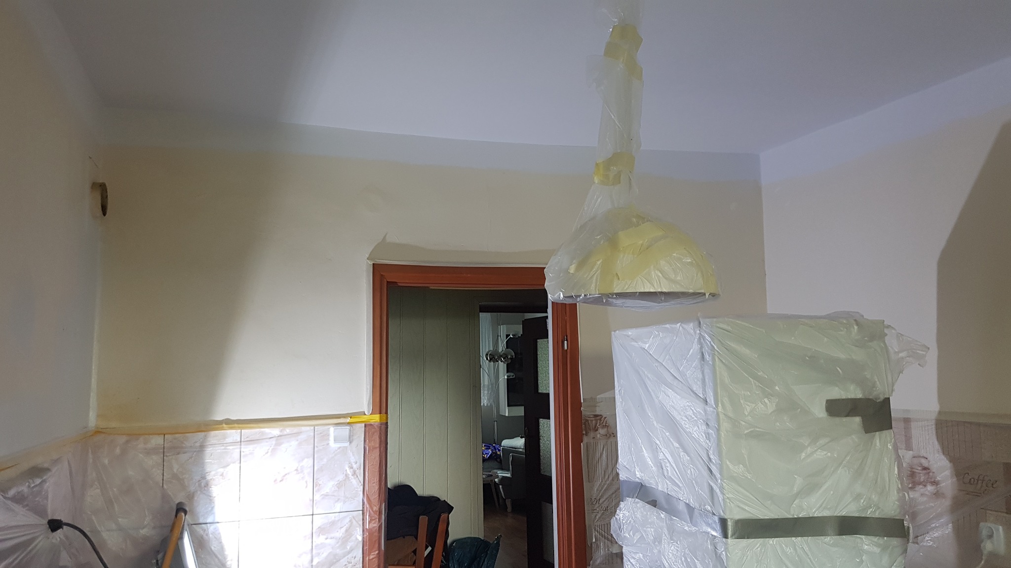 Przygotowanie do odświeżenia ścian w pomieszczeniu: meble i lampa zabezpieczone folią malarską, widoczne świeżo pomalowane górne partie ścian i sufit.