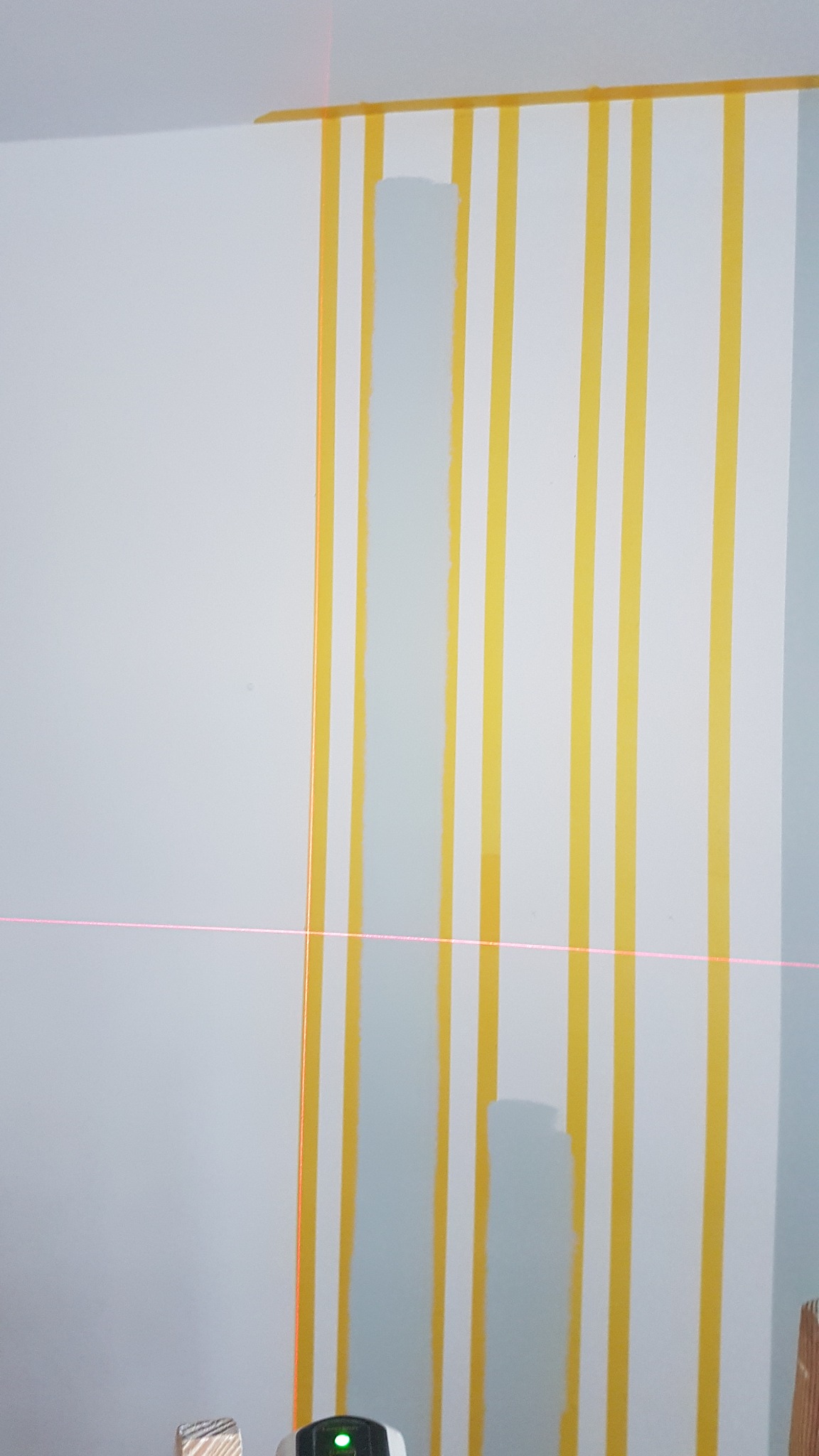 Ściana z pionowymi pasami wyznaczonymi taśmą malarską, częściowo pomalowana na szaro, widoczny laserowy poziomnica.