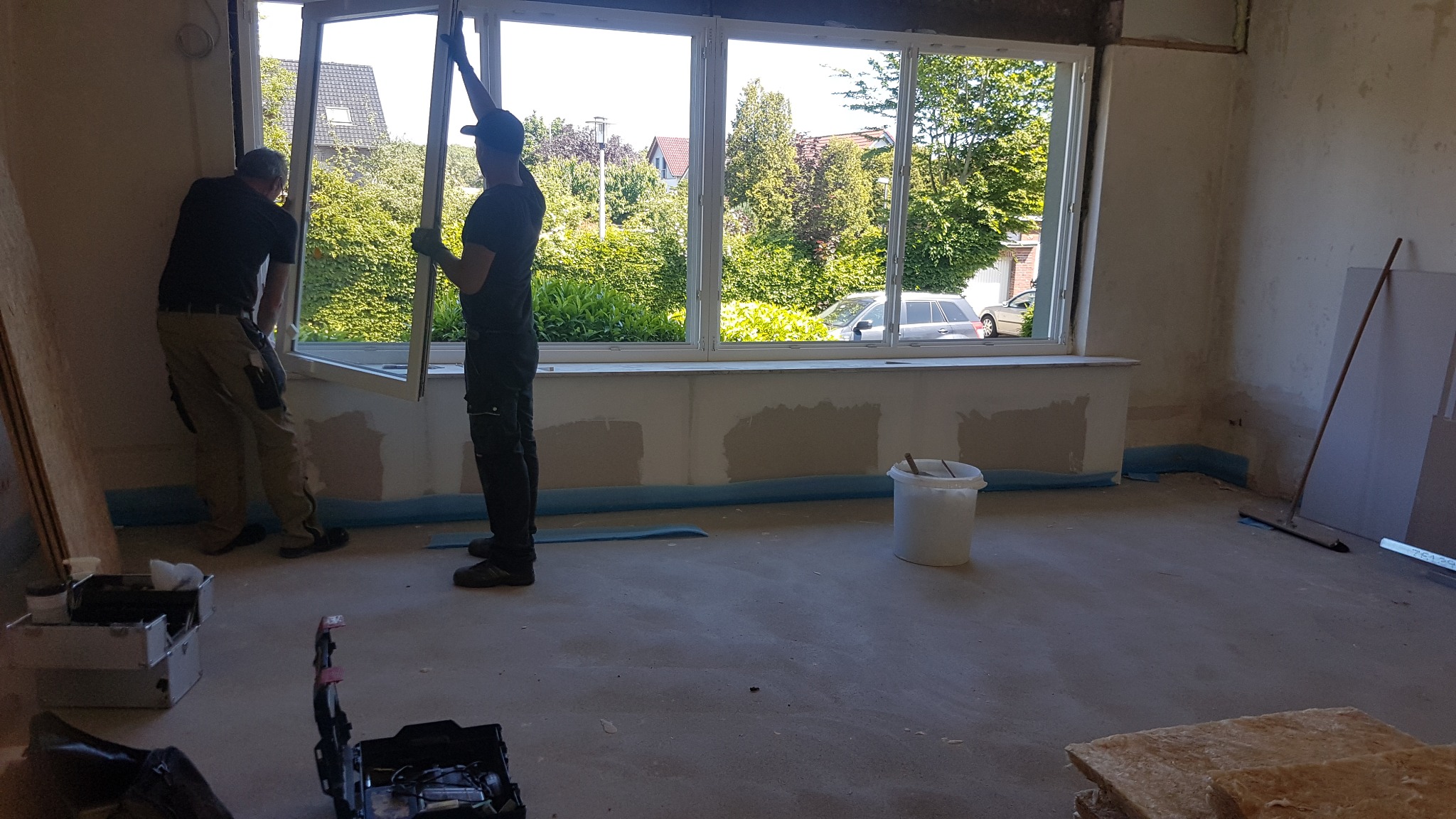 Dwóch monterów instaluje okno panoramiczne w surowym wnętrzu. Widoczne narzędzia, wiadro i płyty gipsowe. Za oknem zieleń i zaparkowany samochód.