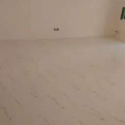 Jasna, błyszcząca podłoga z płytek imitujących marmur w pustym pokoju z dużym oknem. Na ścianie widoczne zielone taśmy malarskie i gniazdka elektryczne.