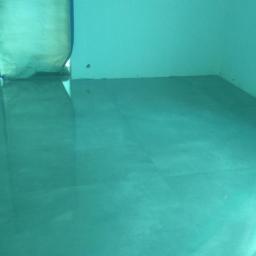 NEST BUDOWNICTWO Karol Flis - Świeżo wylana, samopoziomująca wylewka podłogowa w pustym pomieszczeniu, widoczne kable elektryczne wystające ze ściany.