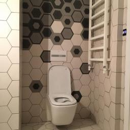 NEST BUDOWNICTWO Karol Flis - Nowoczesna toaleta podwieszana z otwartą klapą, biały grzejnik drabinkowy i heksagonalne płytki w odcieniach szarości i bieli na ścianach i podłodze.