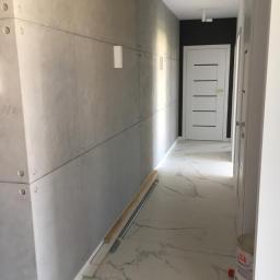 NEST BUDOWNICTWO Karol Flis - Korytarz z wykończonymi ścianami z szarych płyt imitujących beton, marmurowa podłoga, białe drzwi i listwy przypodłogowe, widoczne narzędzia i materiały budowlane w trakcie prac wykończeniowych.