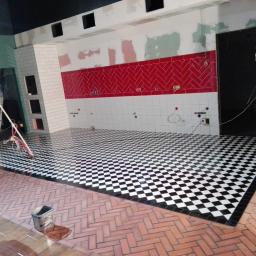 NEST BUDOWNICTWO Karol Flis - Wnętrze w trakcie prac wykończeniowych: ułożona czarno-biała szachownica na podłodze, fragment ściany z czerwonymi płytkami ułożonymi w jodełkę, widoczne przewody i narzędzia, w tle pracownik...