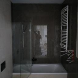 NEST BUDOWNICTWO Karol Flis - Wykończona łazienka z wanną obudowaną jasnymi płytkami, szklanym parawanem nawannowym z czarną armaturą i grzejnikiem drabinkowym. W lustrze odbija się osoba robiąca zdjęcie na statywie.