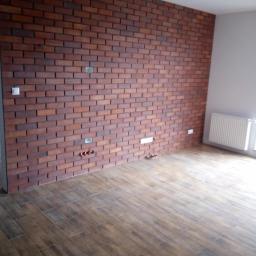 NEST BUDOWNICTWO Karol Flis - Pokój z wykończoną ścianą z cegły klinkierowej w odcieniach brązu i podłogą z płytek imitujących drewno, widoczne gniazdka elektryczne i wyprowadzone rury instalacji wodnej.
