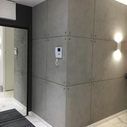 NEST BUDOWNICTWO Karol Flis - Wykończone wnętrze przedpokoju z szarymi panelami ściennymi imitującymi beton, połączonymi metalowymi nitami, biała marmurowa podłoga i czarna rama drzwi.