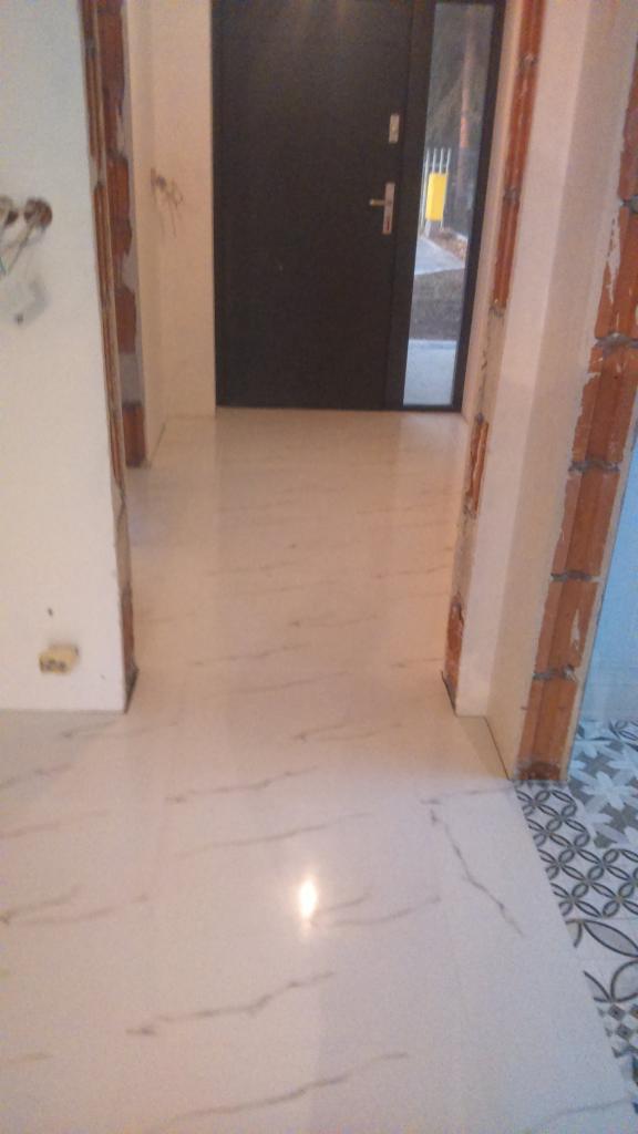 Biała, marmurkowana podłoga z połyskiem w trakcie prac wykończeniowych, widoczne fragmenty surowej cegły i przygotowane do malowania ściany, czarne drzwi wejściowe z przeszkleniem.