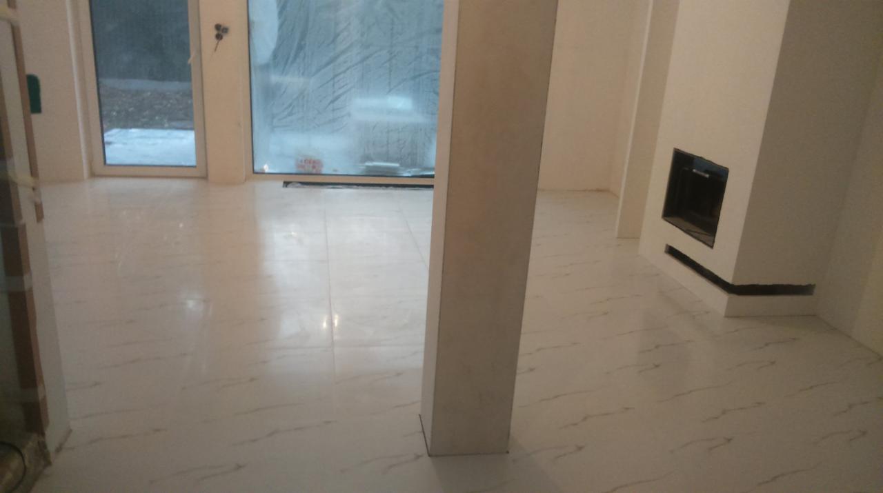 Jasne, błyszczące płytki podłogowe z subtelnym marmurkowym wzorem, świeżo ułożone w przestronnym, jasnym wnętrzu z nowoczesnym kominkiem i dużym oknem.