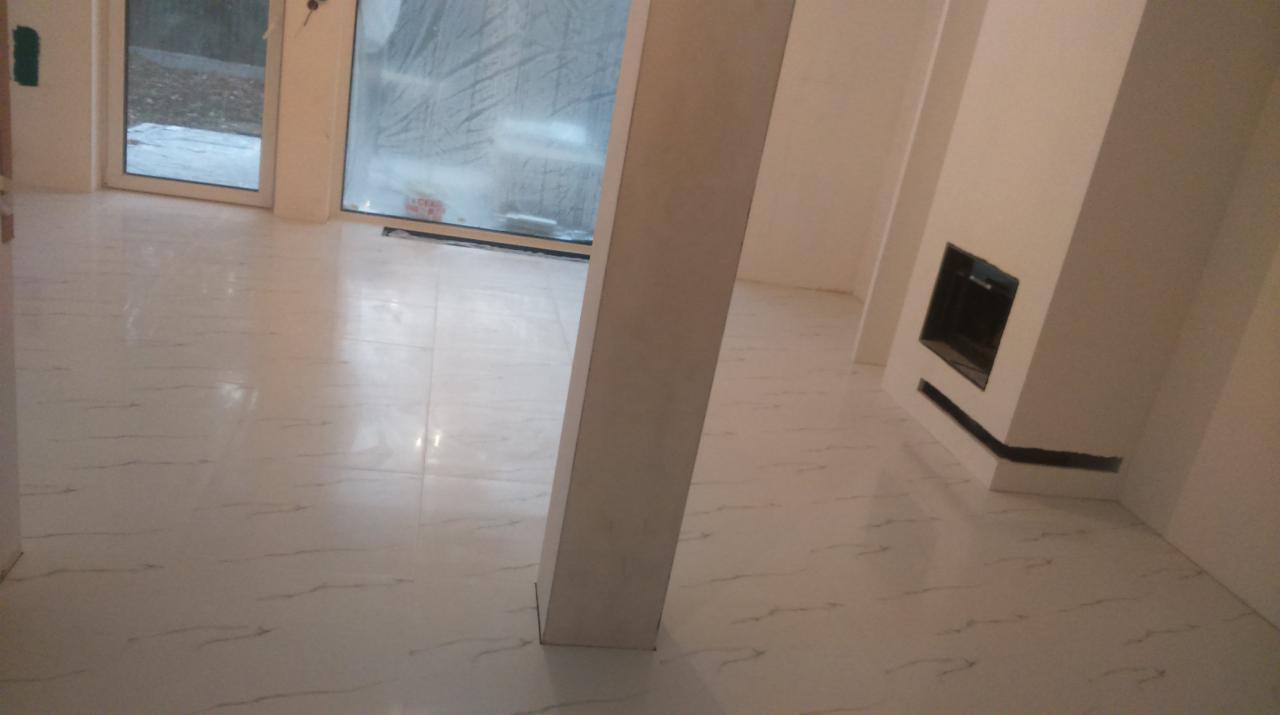 Wykończone wnętrze z jasnymi płytkami podłogowymi imitującymi marmur, kolumną i obudową kominka z czarnym wkładem, widoczne okno z widokiem na zewnątrz.