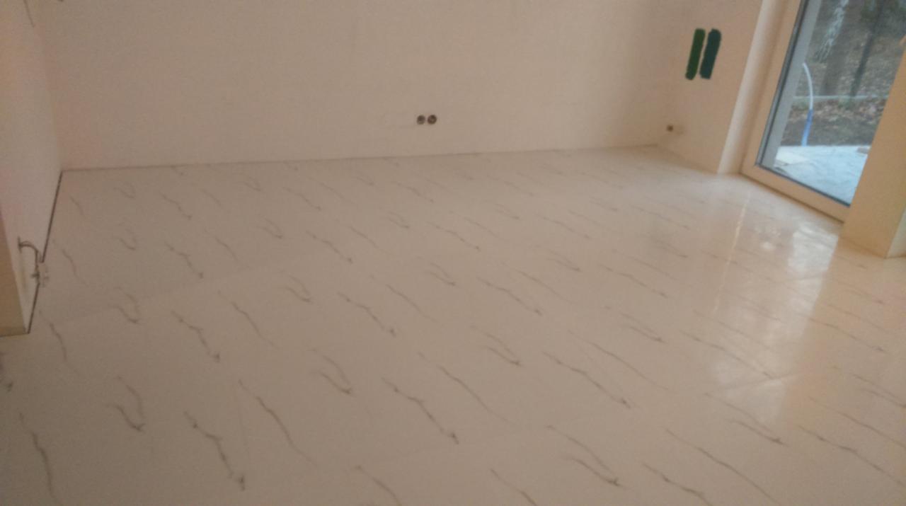 Jasna, błyszcząca podłoga z płytek imitujących marmur w pustym pokoju z dużym oknem. Na ścianie widoczne zielone taśmy malarskie i gniazdka elektryczne.