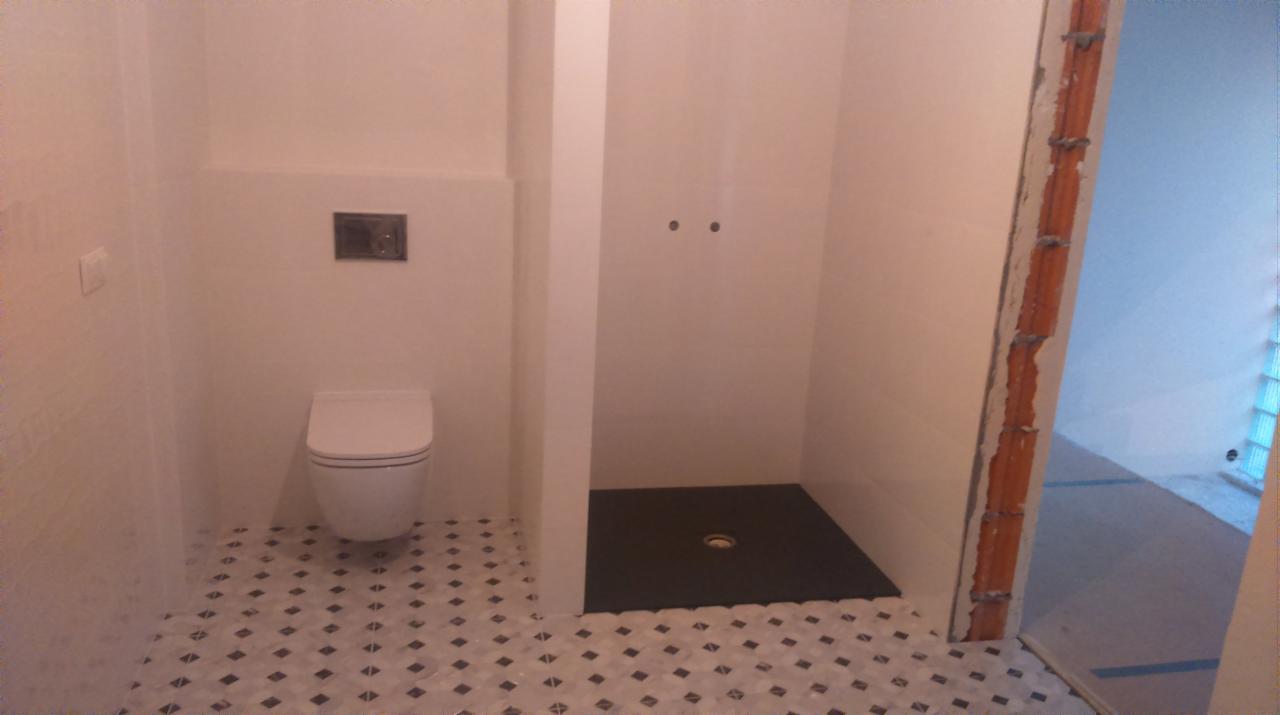 Nowoczesna łazienka z białą ceramiką, kabiną prysznicową typu walk-in z czarnym brodzikiem i wzorzystą podłogą w odcieniach szarości i bieli, widoczny surowy fragment muru przy wejściu.