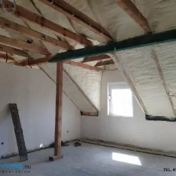 Poddasze domu w trakcie izolacji: widoczne drewniane krokwie i biała pianka izolacyjna na skośnych ścianach, okno dachowe i pionowe okno w ścianie, surowy betonowy podkład podłogowy.