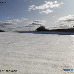 Biała, szorstka powierzchnia dachu po natrysku izolacji, widoczna antena telewizyjna, logo firmy natryskowe.eu w rogu.