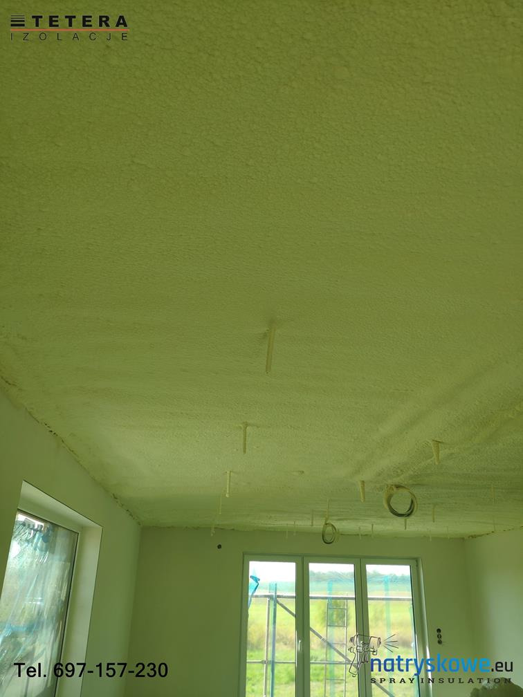 Poddasze po natrysku pianki izolacyjnej, widoczne wystające elementy montażowe i niedokładności aplikacji przy ścianach, logo firmy w rogu.