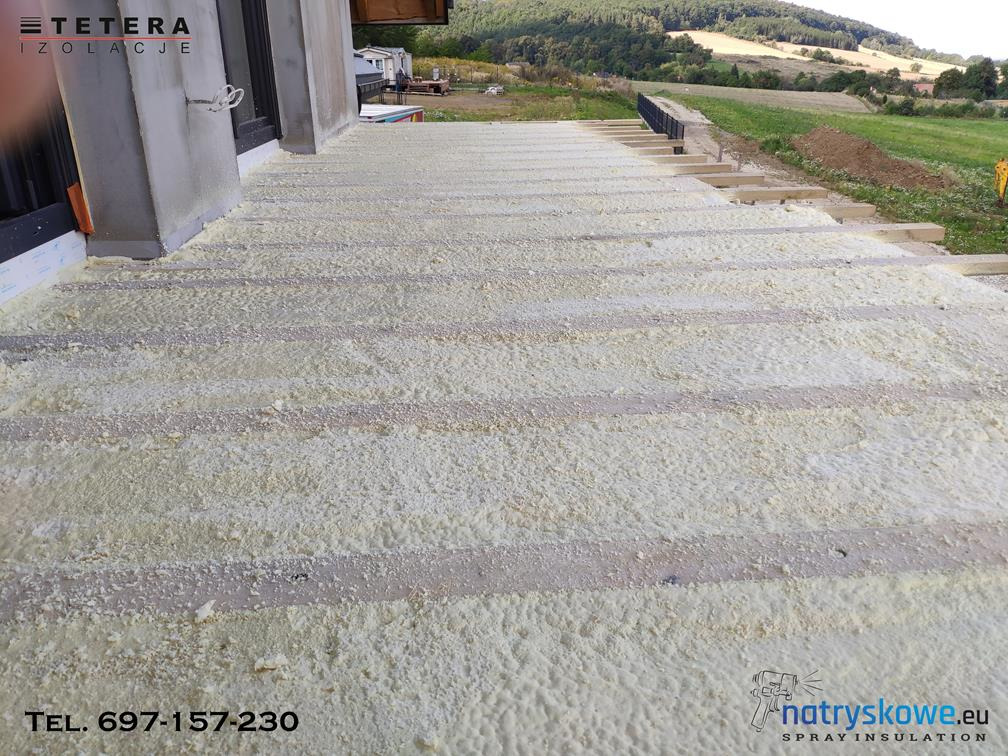 Świeżo natryśnięta, jasna pianka izolacyjna wypełniająca przestrzeń między legarami pod przyszły taras, widok z góry na nierówną strukturę piany i krajobraz w tle.