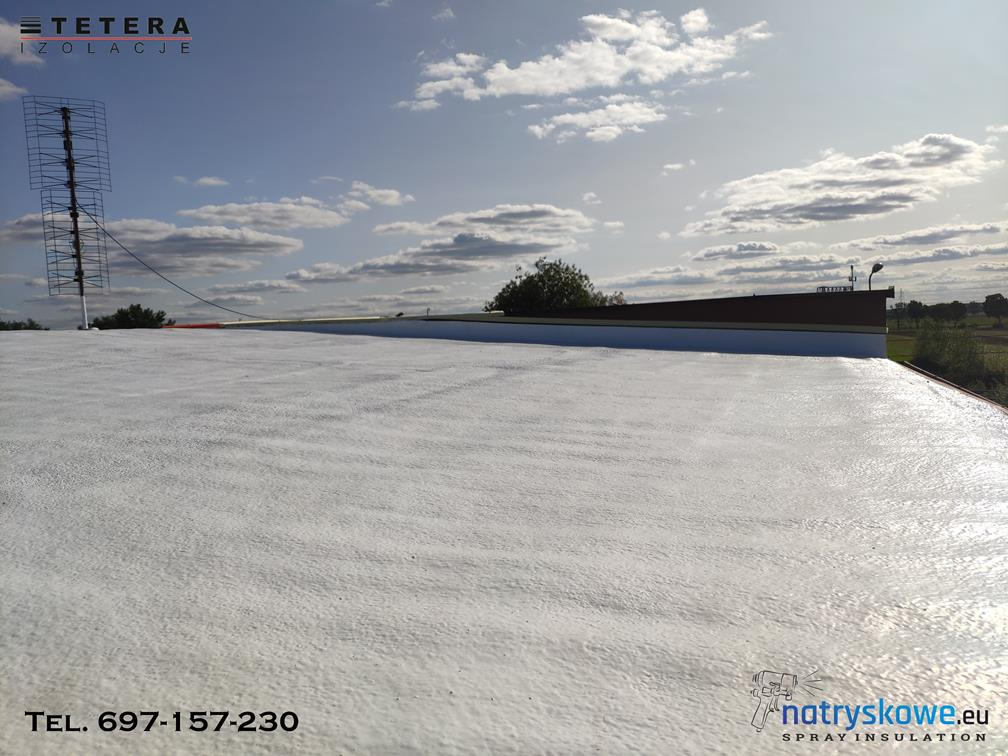 Biała, szorstka powierzchnia dachu po natrysku izolacji, widoczna antena telewizyjna, logo firmy natryskowe.eu w rogu.