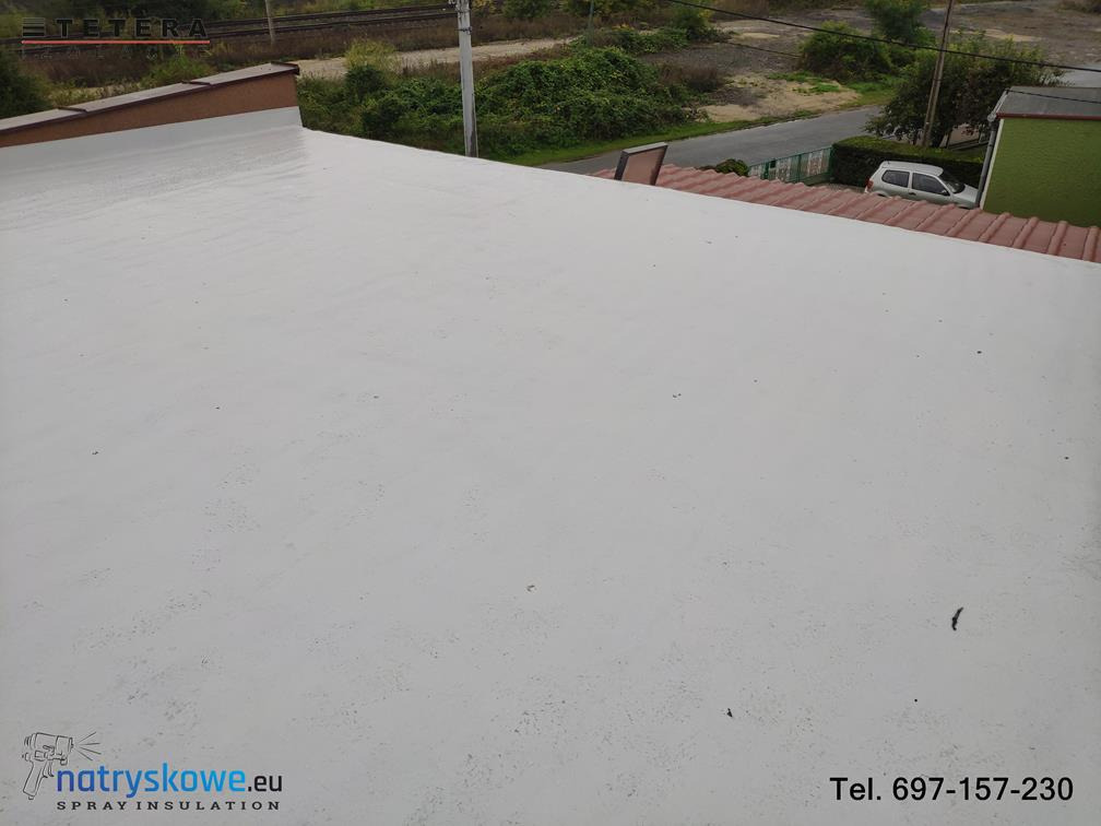Wykończona, jasnoszara, natryskowa izolacja dachu płaskiego z widocznym logo firmy natryskowe.eu i numerem telefonu w prawym dolnym rogu, w tle roślinność i torowisko kolejowe.