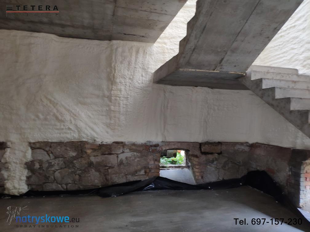Ściana piwnicy z kamienną podstawą po natrysku białej piany izolacyjnej, widoczny betonowy strop i schody, małe okienko z widokiem na zieleń.